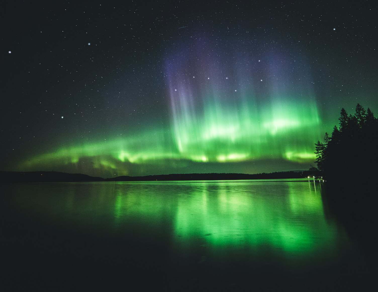 A aurora boreal em Kuopio com tons de verde e roxo ilumina um céu estrelado sobre um lago calmo cercado por árvores. Representa eSIM Finlândia.