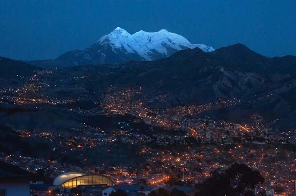 Paisagem urbana da cidade La Paz à noite com ruas iluminadas, tendo como pano de fundo montanhas cobertas de neve sob um céu claro e escuro. Representa eSIM Bolívia.