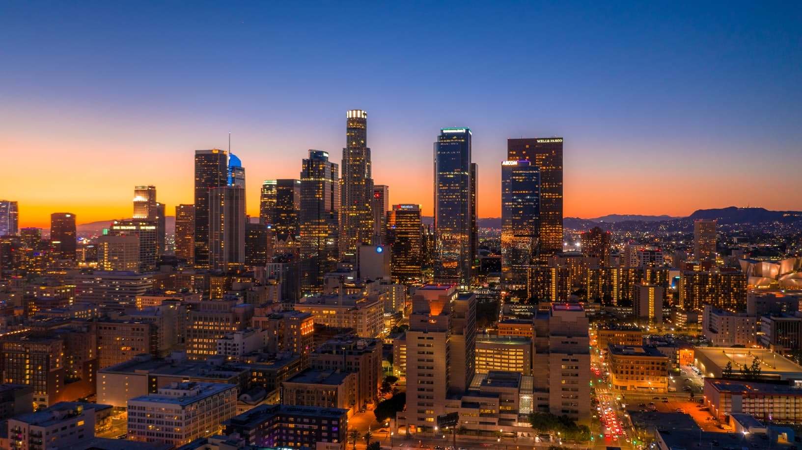Vista aérea do horizonte da cidade de Los Angeles ao anoitecer, com edifícios altos iluminados contra um céu laranja e azul. Representa eSIM Los Angeles.