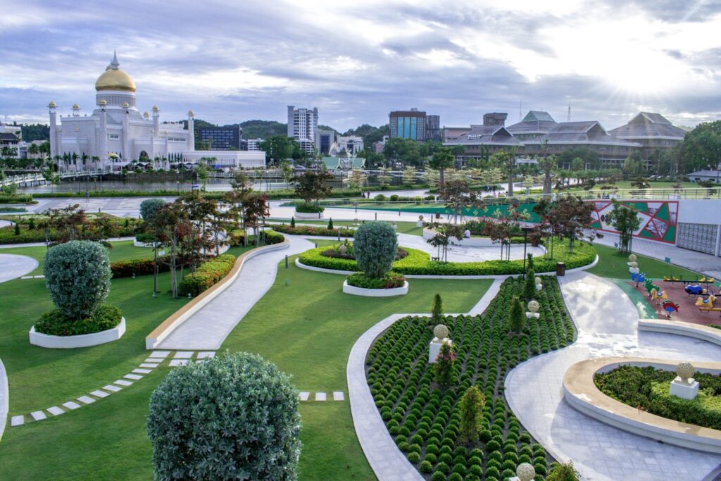Um parque paisagístico com caminhos e áreas verdes, com uma mesquita abobadada e prédios da cidade ao fundo, sob um céu nublado, em Brunei.