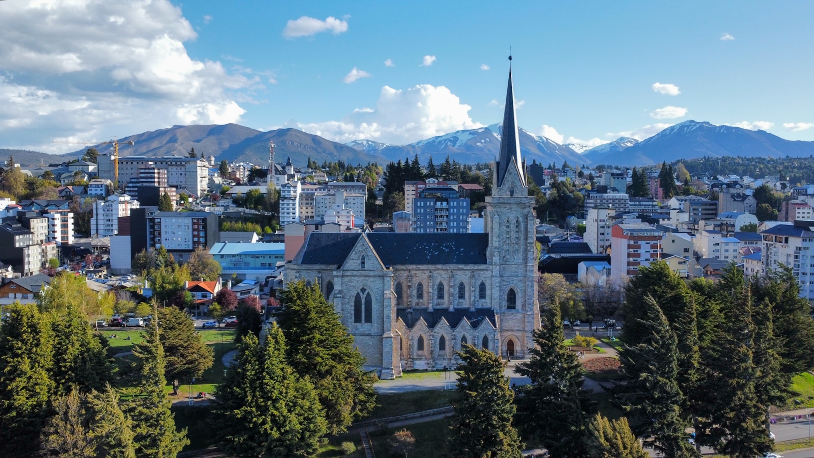 Uma igreja de pedra com uma torre alta é cercada por árvores em uma paisagem urbana, com montanhas ao fundo sob um céu parcialmente nublado, em Bariloche. Representa eSIM Bariloche.