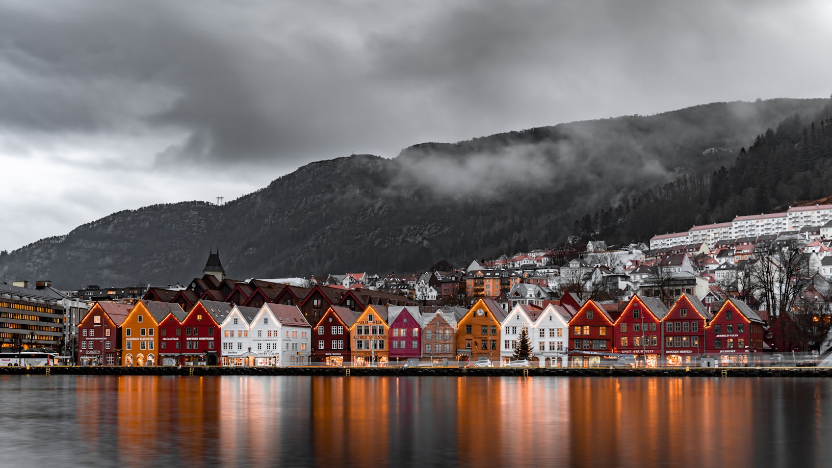 Casas coloridas se alinham à beira-mar em Bryggen, tendo como pano de fundo montanhas enevoadas e um céu nublado, com reflexos em águas calmas. Representa eSIM Noruega.