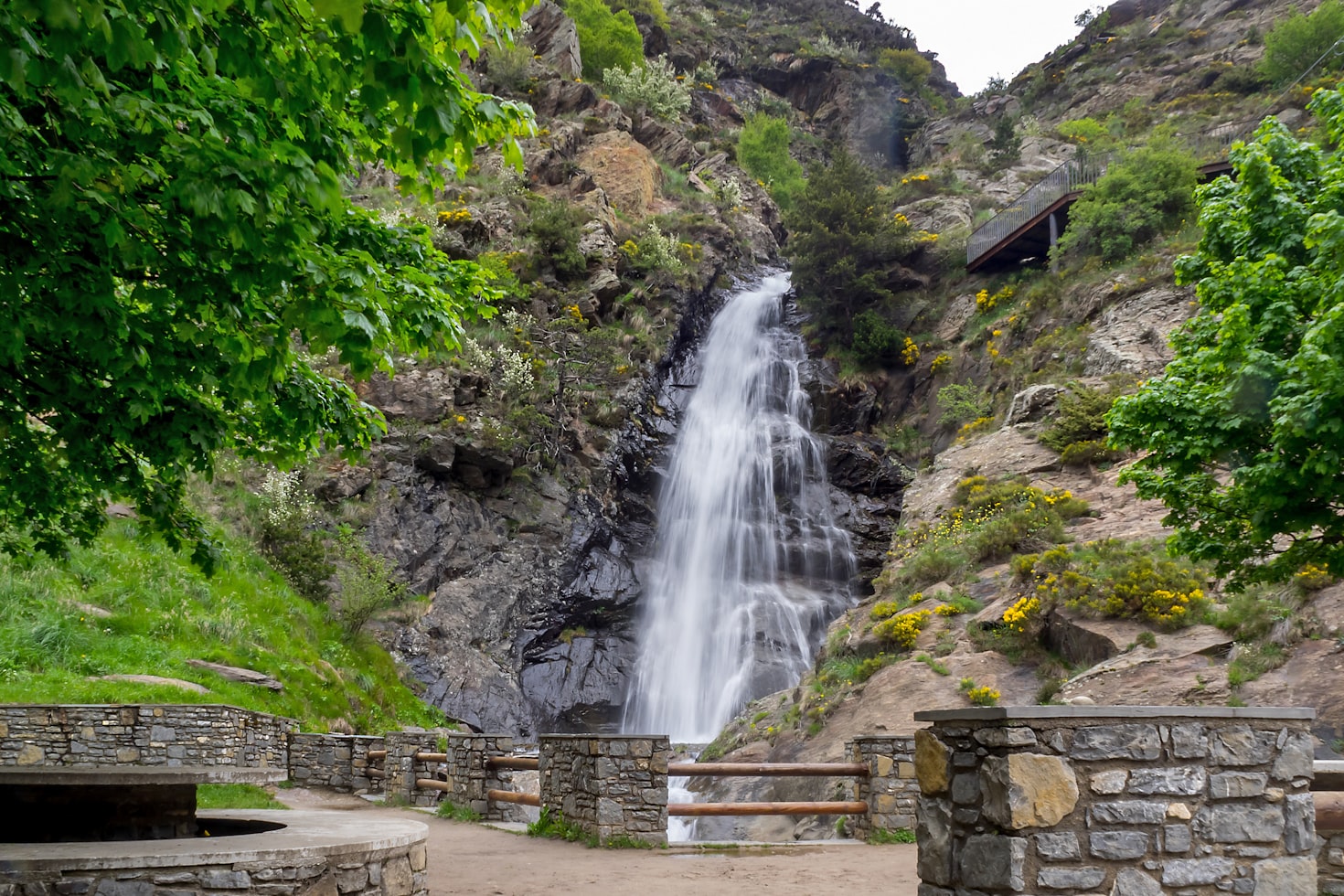Cachoeira caindo em cascata sobre um penhasco rochoso cercado por árvores verdes, com uma área de observação de pedra e grades em primeiro plano.