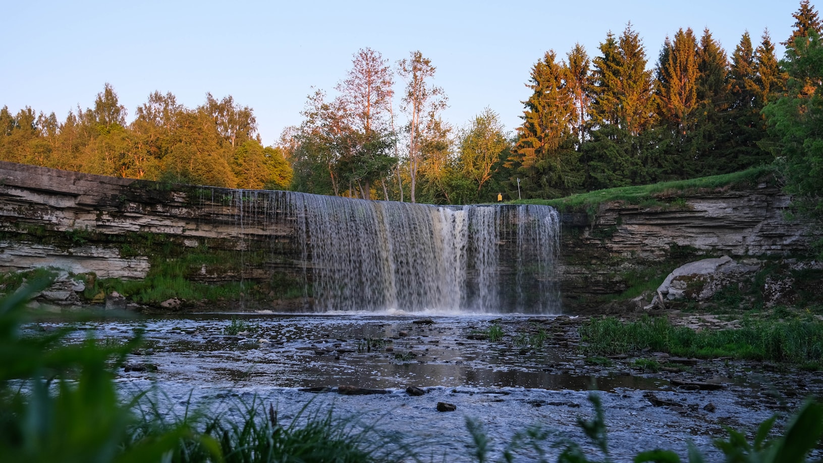 Uma cachoeira desce sobre um penhasco rochoso cercado por árvores e vegetação, com um rio calmo fluindo abaixo dela, na Estônia. Representa eSIM Estônia.