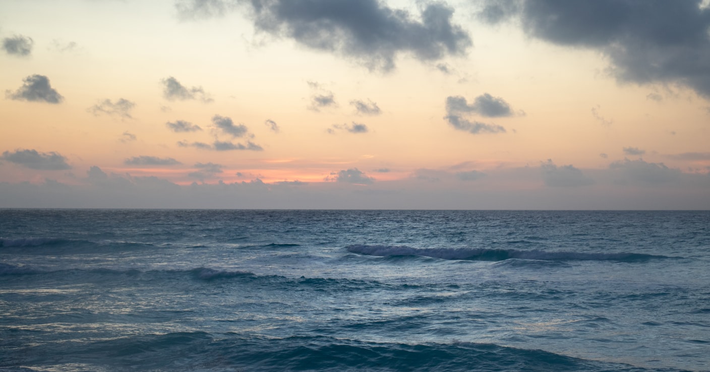 Ondas calmas do oceano sob um céu nublado ao pôr do sol, com cores suaves de laranja e rosa no horizonte,em Cancun.Representa eSIM Cancun.