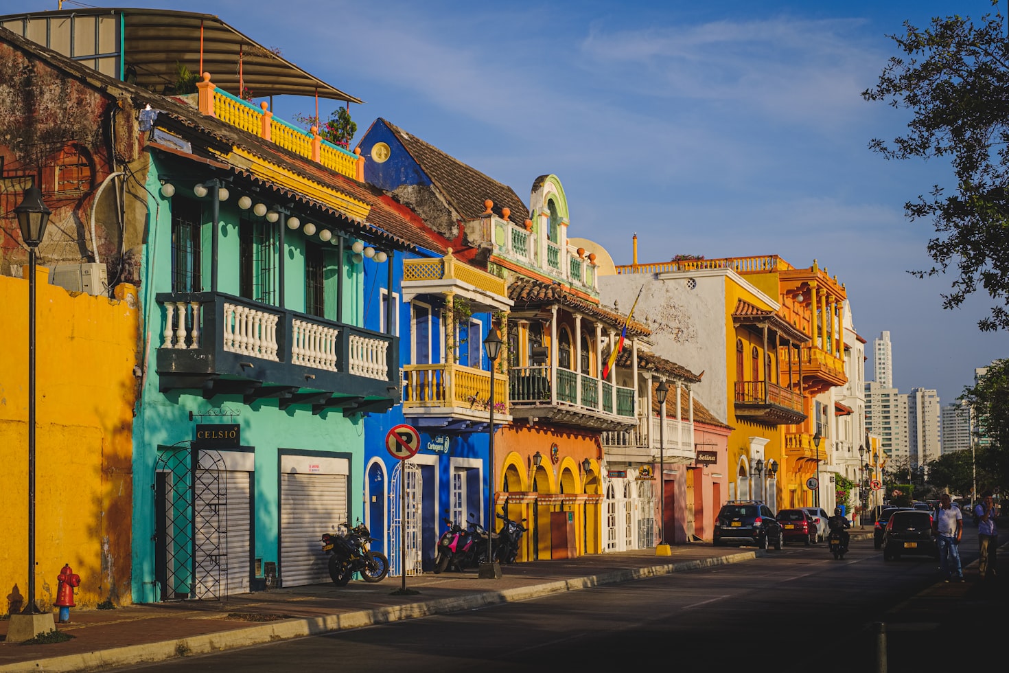 Uma rua ladeada em Cartagena por edifícios vibrantes e coloridos em uma área histórica sob um céu azul. Carros e bicicletas estacionados ao longo da rua. Representa eSIM Cartagena.