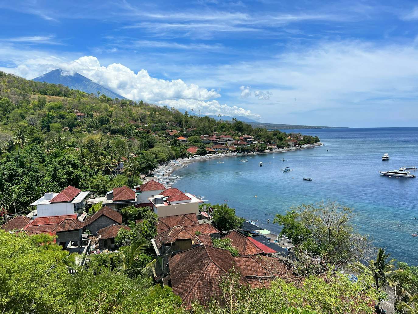 Vista de uma vila costeira com casas de telhados vermelhos, colinas verdes, uma montanha ao longe e barcos em águas azuis, em Amed Beach. Representa eSIM Indonésia.