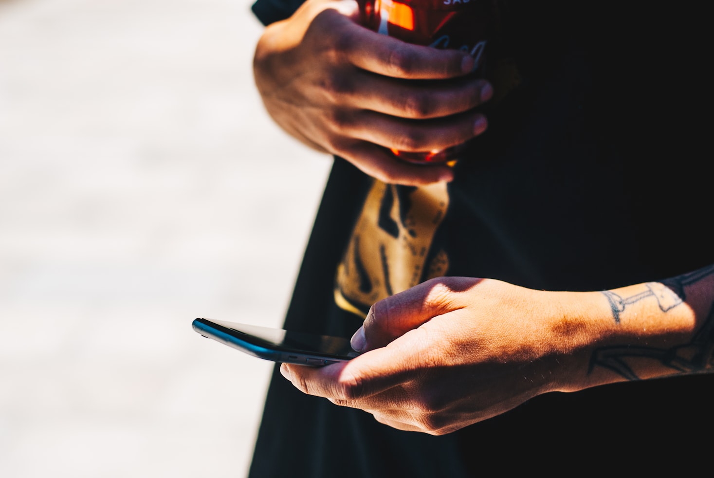 Pessoa segurando um smartphone em uma mão e uma lata de bebida na outra, com uma tatuagem visível no braço.