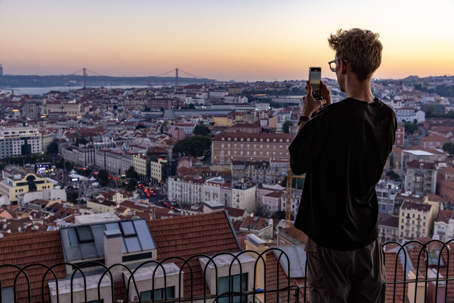 Uma pessoa tira uma foto com um smartphone observando uma paisagem urbana ao pôr do sol, com um rio e uma ponte ao fundo. Representa chip internacional.