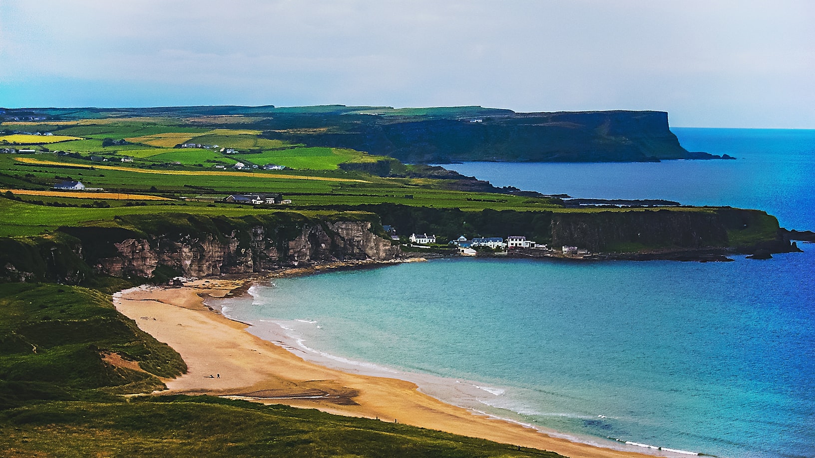 Paisagem costeira com praia de areia, falésias verdes e casas espalhadas à beira-mar sob um céu parcialmente nublado.