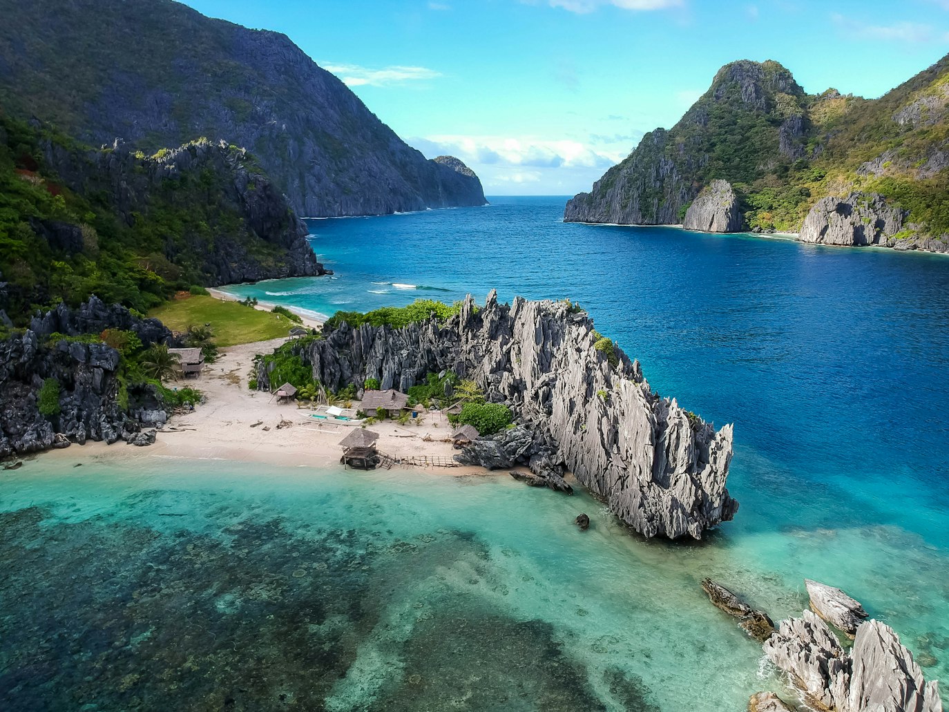Uma formação rochosa de calcário separa uma praia arenosa de águas azul-turquesa, com colinas e montanhas verdes ao fundo, em El Nido. Representa eSIM Filipinas.