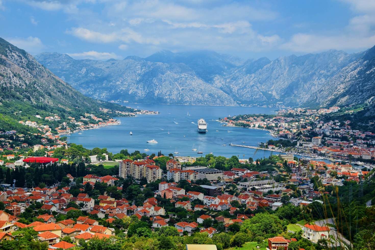 Vista panorâmica de Kotor com edifícios com telhados vermelhos, um navio de cruzeiro e montanhas ao redor de uma baía sob um céu parcialmente nublado. Representa eSIM Montenegro.