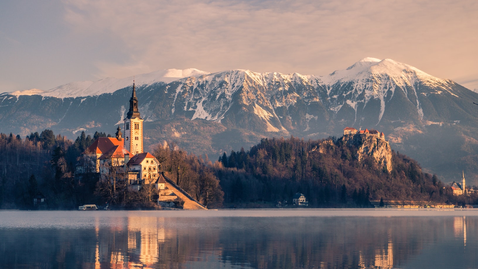 Igreja em uma ilha em um lago Bled com montanhas cobertas de neve e um céu parcialmente nublado ao fundo, refletido na água. Representa eSIM Eslovênia.