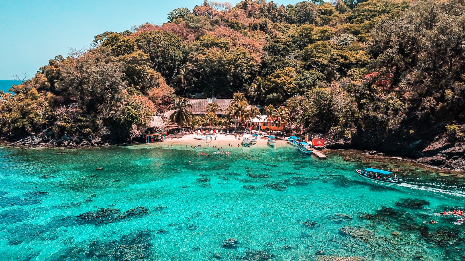 Uma pequena praia tropical com águas cristalinas azul-turquesa, cercada por árvores densas, barcos e vários edifícios na costa em Cayos Cochinos. Representa eSIM Honduras.
