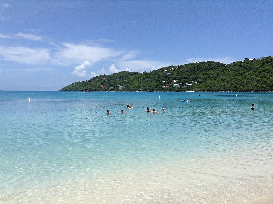 Pessoas nadam em águas rasas e claras perto de uma praia arenosa com colinas verdes e céu azul ao fundo, nas Ilhas Virgens Americanas. Representa eSIM Ilhas Virgens Americanas.