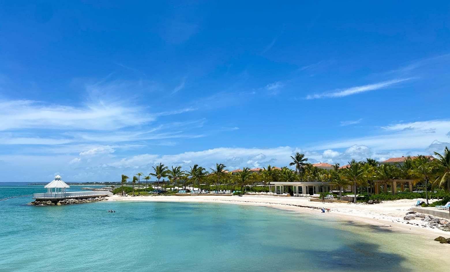 Uma praia tropical com águas cristalinas, palmeiras, um mirante branco em um píer e prédios de resort sob um céu azul, Punta Cana. Representa eSIM Punta Cana.
