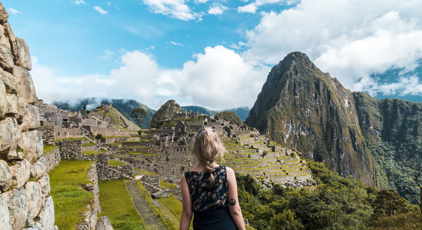 Pessoa em pé em um caminho com vista para as ruínas antigas de Machu Picchu, com montanhas e um céu nublado ao fundo.