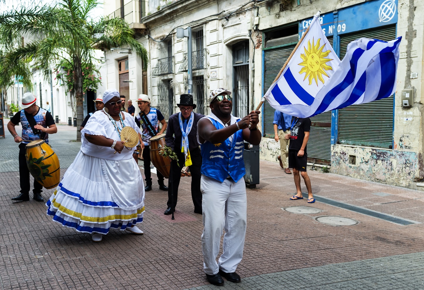 Um grupo de pessoas em trajes festivos desfila por uma rua, com uma pessoa agitando a bandeira uruguaia na frente, em . Representa e SIM Montevideo.