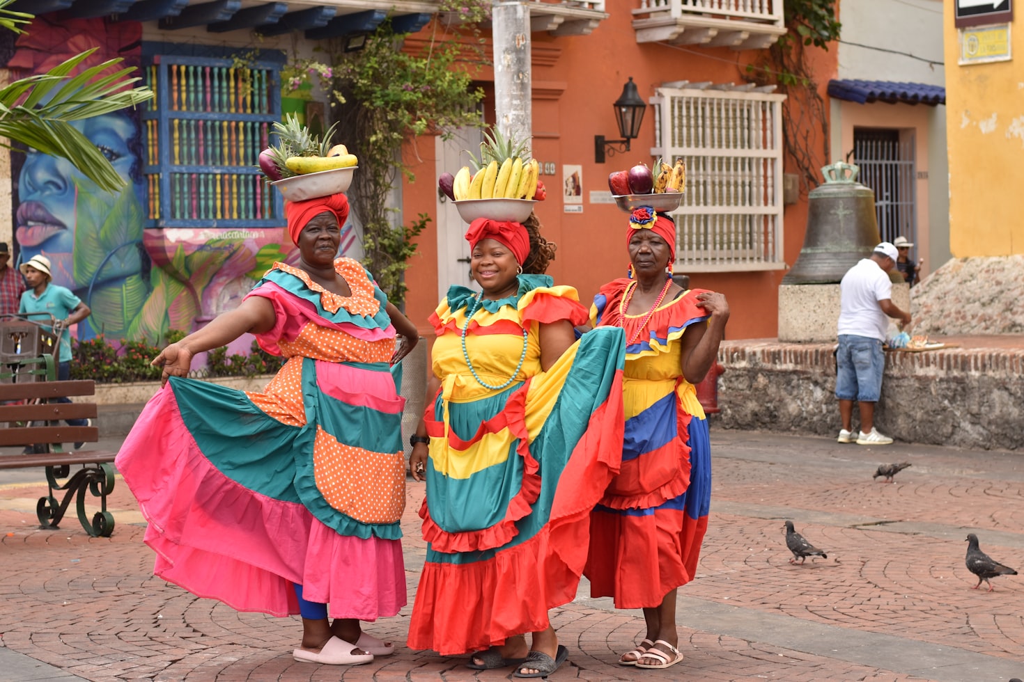 Três mulheres em vestidos coloridos e lenços na cabeça em Cartagena carregam cestas de frutas na cabeça, em um cenário de rua vibrante. Representa eSIM Cartagena.