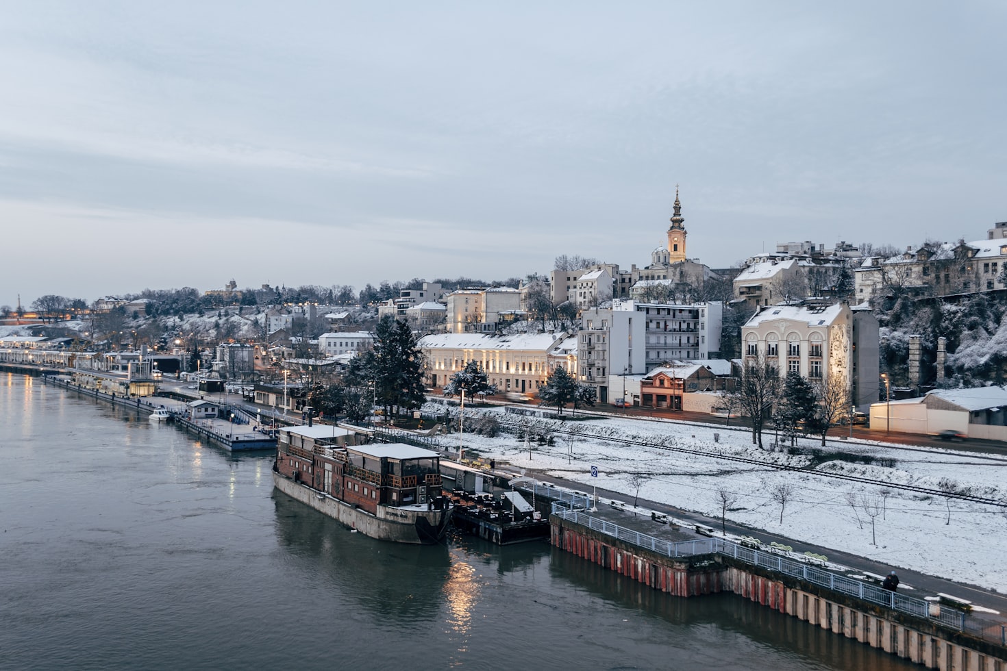 Cidade ribeirinha de Belgrade coberta de neve com barcos atracados no rio. Prédios e uma igreja com uma torre alta alinham a colina. Céu nublado. Representa eSIM Sérvia.