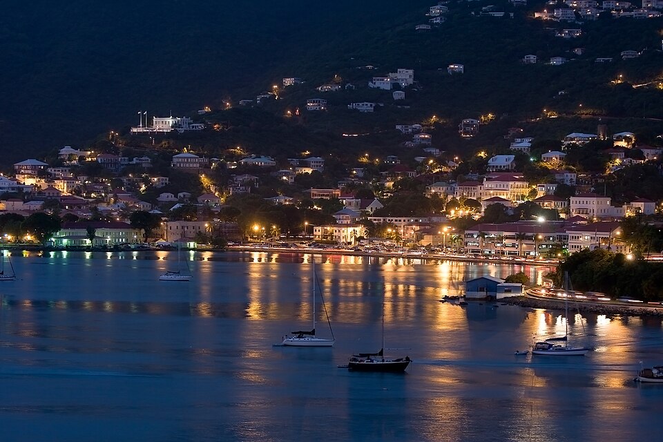 Cidade costeira ao anoitecer com edifícios iluminados, casas nas encostas e barcos ancorados em águas calmas refletindo luzes, nas Ilhas Virgens Americanas. Representa eSIM Ilhas Virgens Americanas.