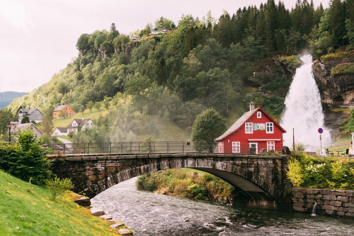 Uma ponte de pedra cruza um rio com uma casa vermelha em Steinsdalsfossen, uma cachoeira e uma encosta arborizada ao fundo, sob um céu nublado. Representa eSIM Noruega.