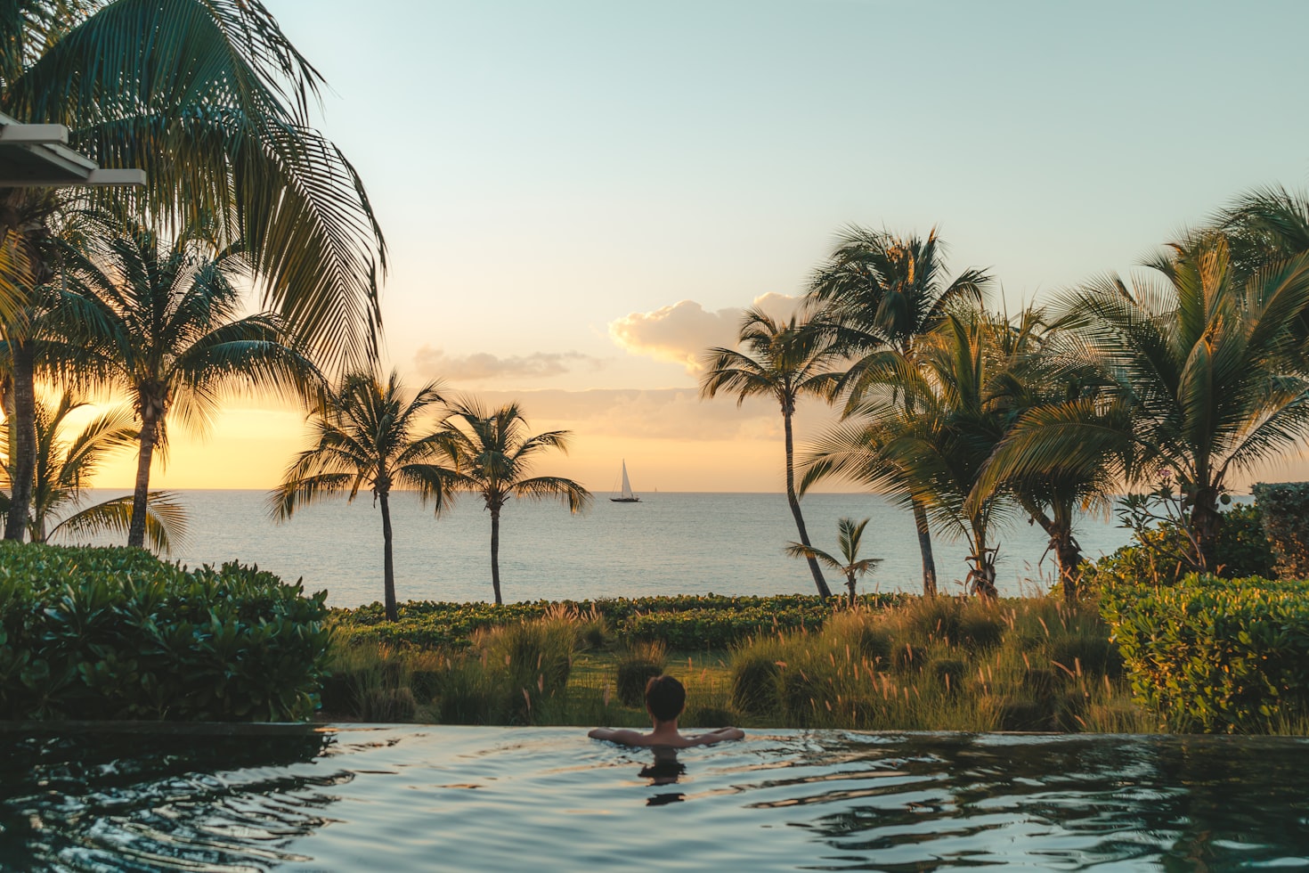 Pessoa em uma piscina com vista para o oceano ao pôr do sol, cercada por palmeiras, com um veleiro ao longe, em Anguilla. Representa eSIM Anguilla.