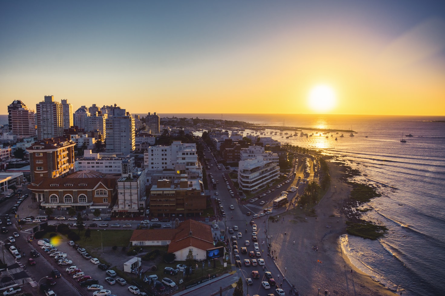Uma cidade costeira em Punta del Este ao pôr do sol com prédios altos, uma estrada movimentada e o oceano à direita, refletindo a luz dourada do sol. no Uruguai. Representa eSIM Uruguai.