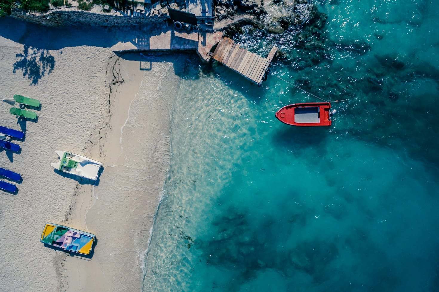 Vista aérea de uma praia de areia de Ksamil com três barcos na costa e um barco vermelho em águas azuis cristalinas perto de um cais de madeira. Representa eSIM Albânia.