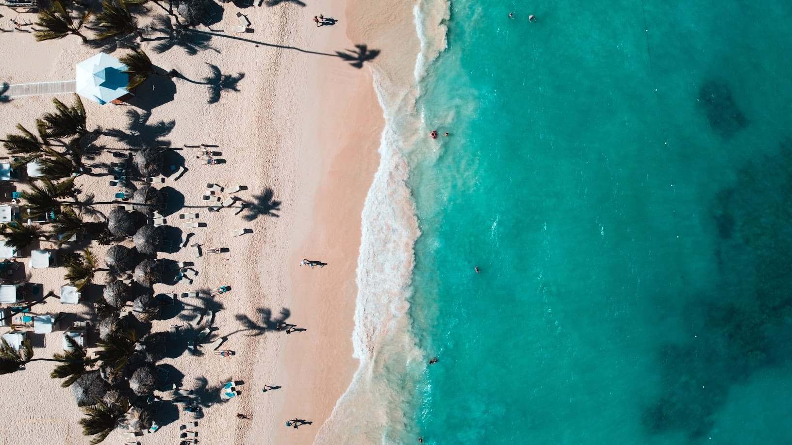 Vista aérea de uma praia de areia com guarda-sóis de palha, pessoas, palmeiras e águas cristalinas azul-turquesa.