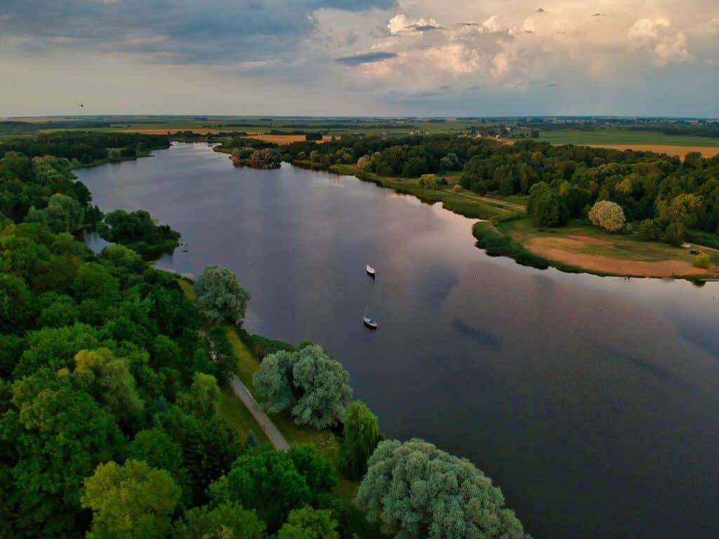 Um rio largo serpenteia por entre árvores verdes e campos sob um céu parcialmente nublado, com dois barcos visíveis na água, em Belarus.