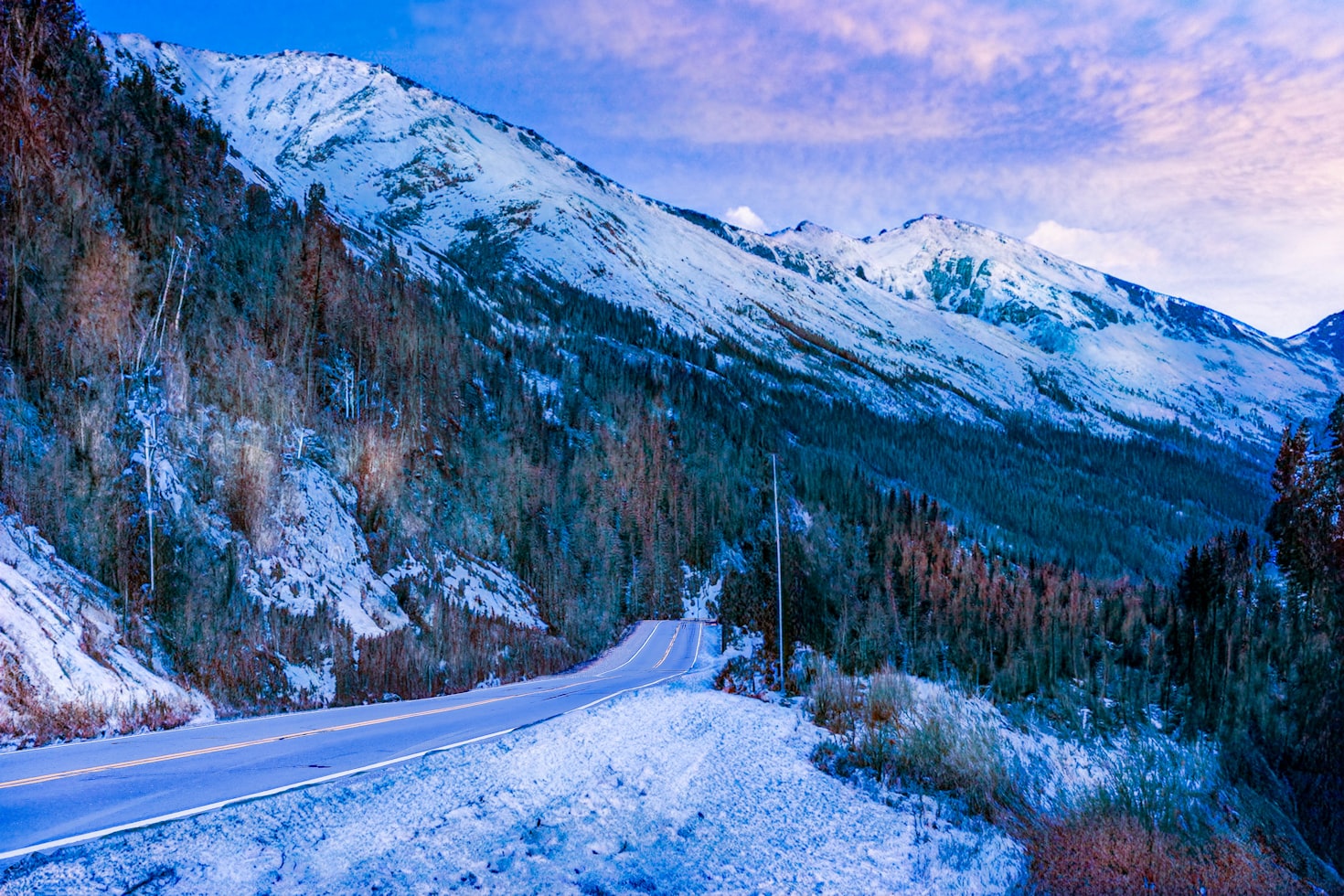 Uma estrada coberta de neve serpenteia por um vale florestado com montanhas ao fundo e um céu parcialmente nublado, em Bariloche. Representa eSIM Bariloche.