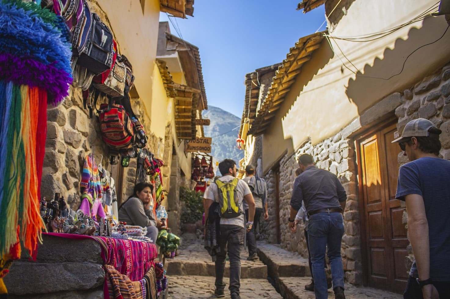 Rua estreita de paralelepípedos em Cusco com turistas caminhando, tecidos coloridos e souvenirs expostos à esquerda e muros de pedra alinhados em ambos os lados. Representa eSIM Cusco.