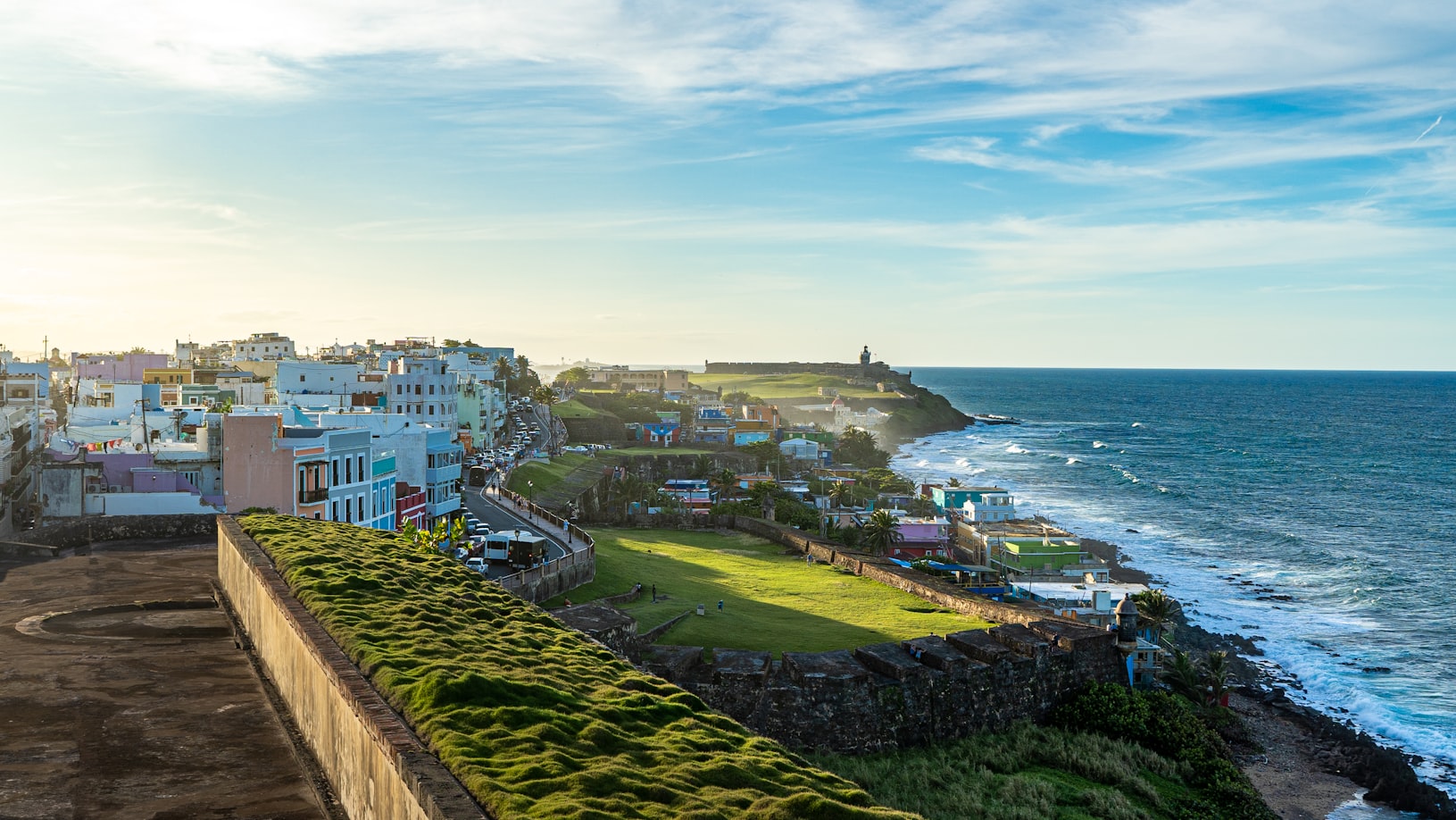 Paisagem urbana costeira com edifícios coloridos, gramados verdes e um farol à beira-mar sob um céu parcialmente nublado, em San Juan. Representa eSIM Porto Rico.