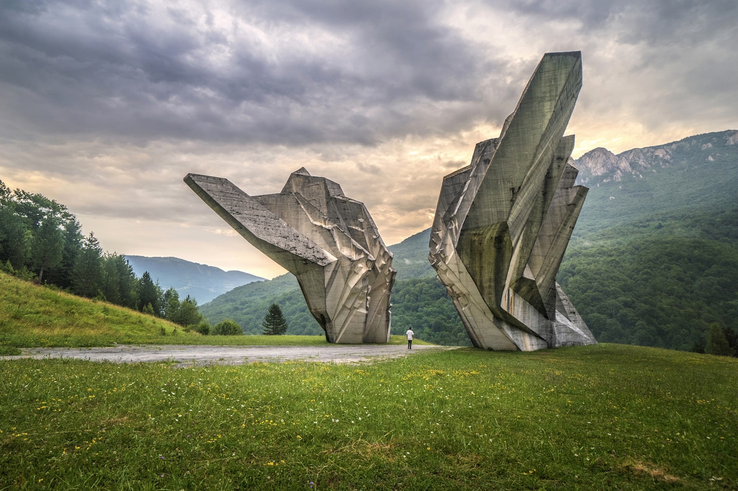 Monumento de concreto com bordas irregulares em uma paisagem gramada, árvores e montanhas ao fundo sob um céu nublado, em Tjentiste War Memorial. Representa eSIM Bósnia e Herzegovina.