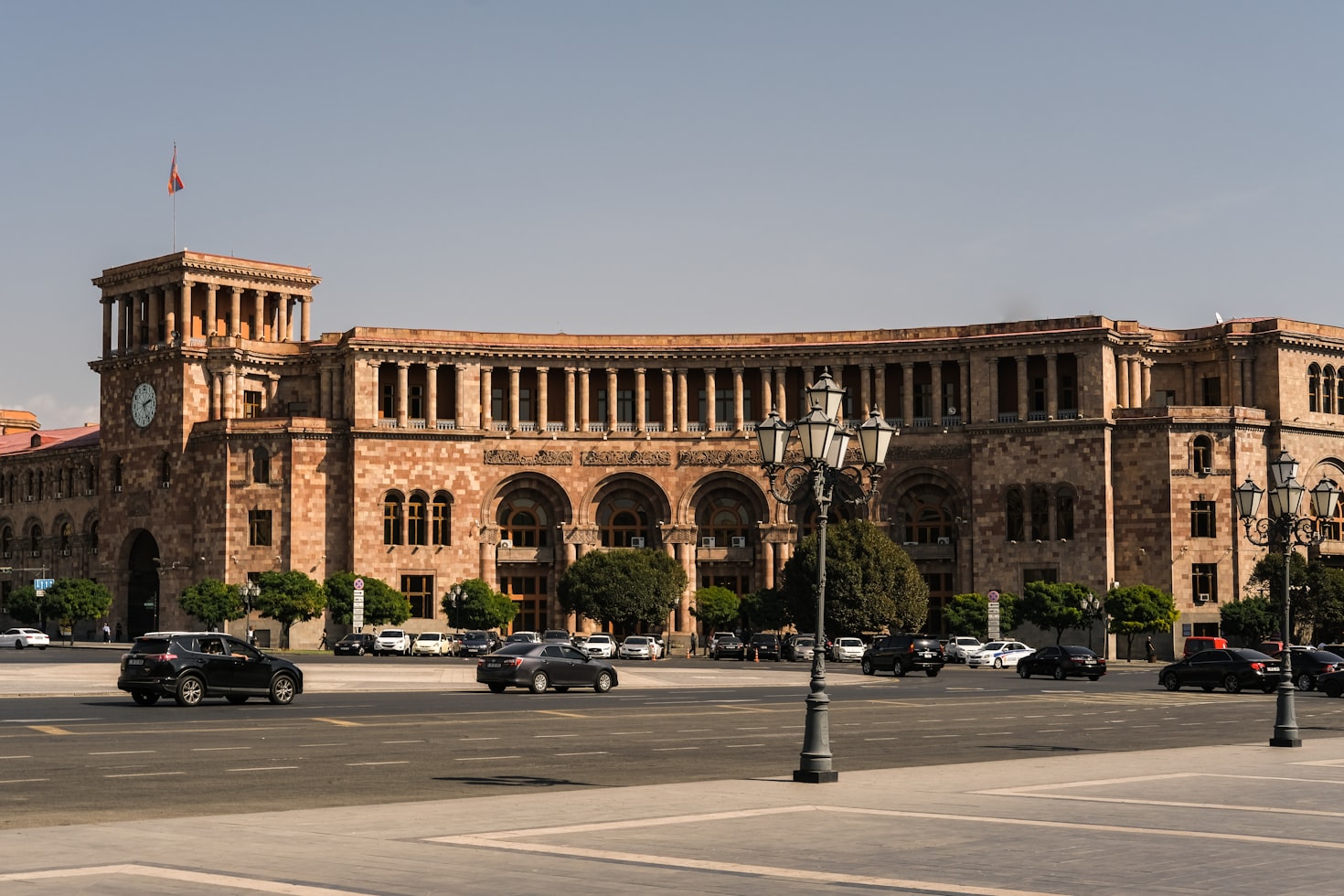 Um grande edifício governamental de pedra com arcos e uma torre do relógio fica ao lado de uma rua com carros e árvores na frente, em Yerevan. Representa eSIM Armênia.