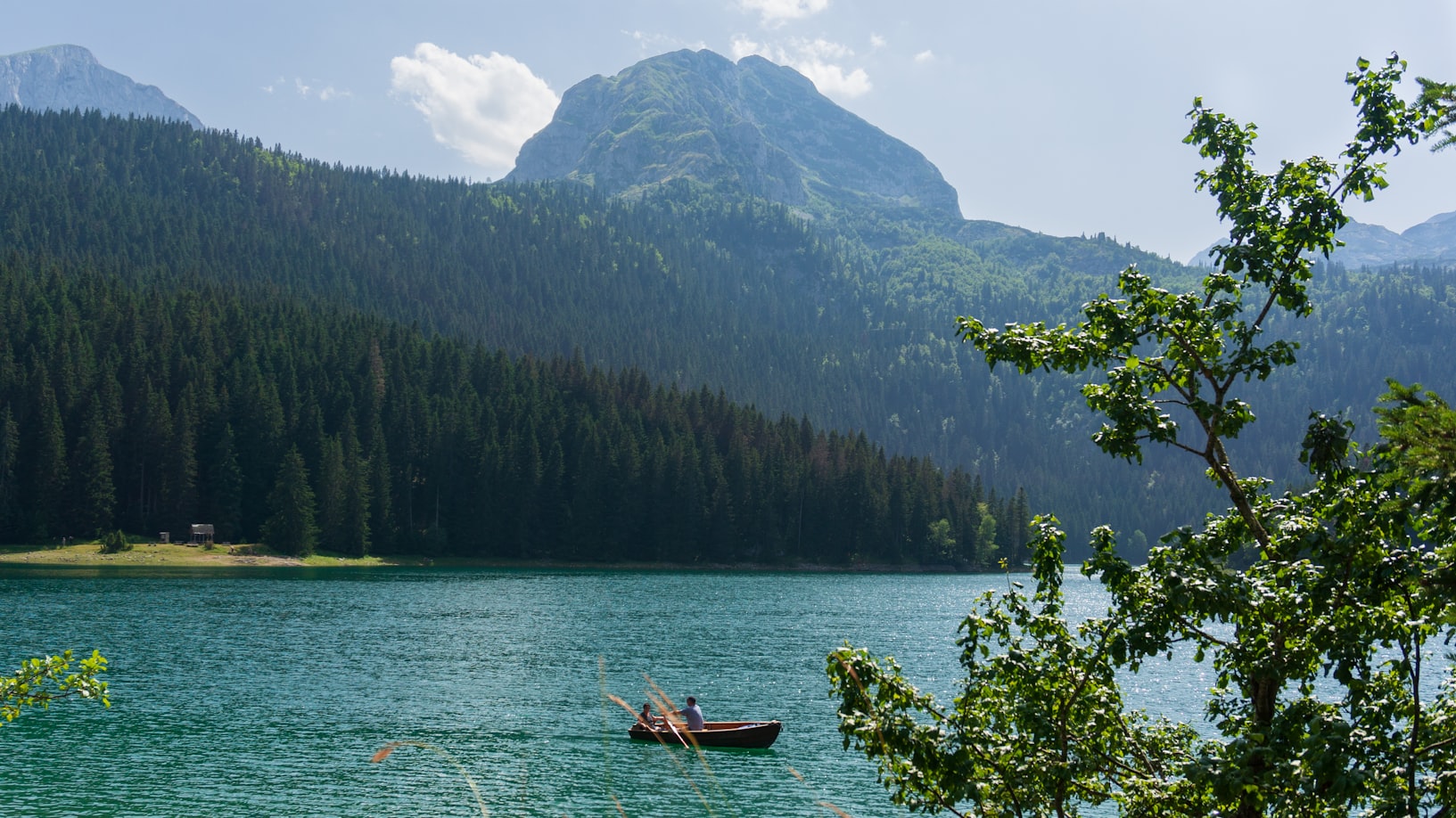 Um pequeno barco em Žabljak, com um lago cercado por colinas arborizadas e uma montanha ao fundo, com uma árvore frondosa em primeiro plano. Representa eSIM Montenegro.