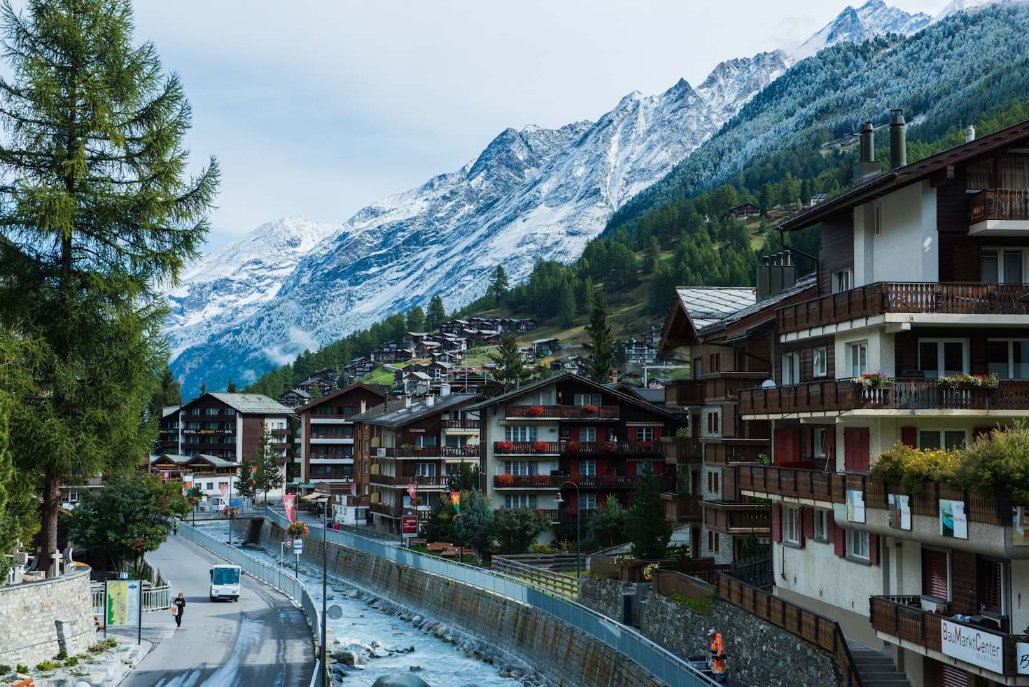 Cidade montanhosa de Zermatt com picos cobertos de neve, prédios de madeira, rio e uma rua arborizada. Ônibus e pedestres visíveis. Representa eSIM Suíça.