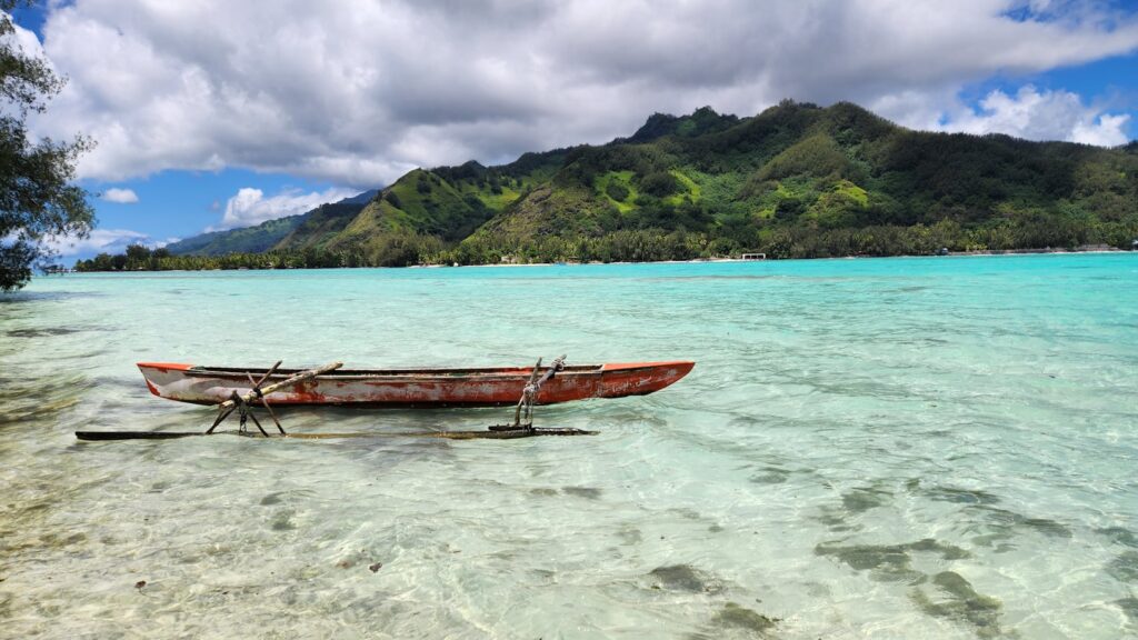 Uma canoa de madeira vermelha flutua em águas rasas e claras, com uma montanha verde e um céu nublado ao fundo, em Moorea. Representa eSIM Polinésia Francesa.
