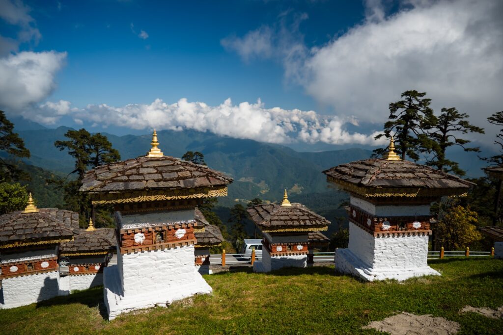 Fileiras de stupas brancas e douradas com montanhas e nuvens ao fundo sob um céu azul.