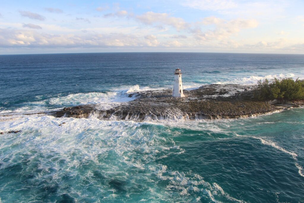 Um farol fica em uma ilha rochosa cercada por ondas do oceano, com árvores à direita e um céu parcialmente nublado, em Bahamas. Representa eSIM Bahamas.