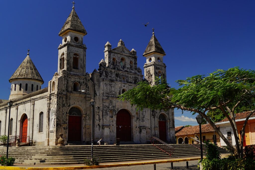 Igreja histórica de pedra com torres, portas vermelhas em arco e degraus largos sob um céu azul claro e com árvores verdes por perto, em Granada. Representa eSIM Nicarágua.