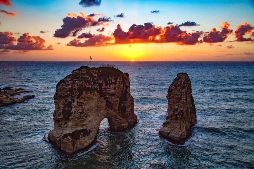 Duas grandes rochas marinhas emergem do oceano com o sol se pondo no horizonte e nuvens no céu, em Beirute.