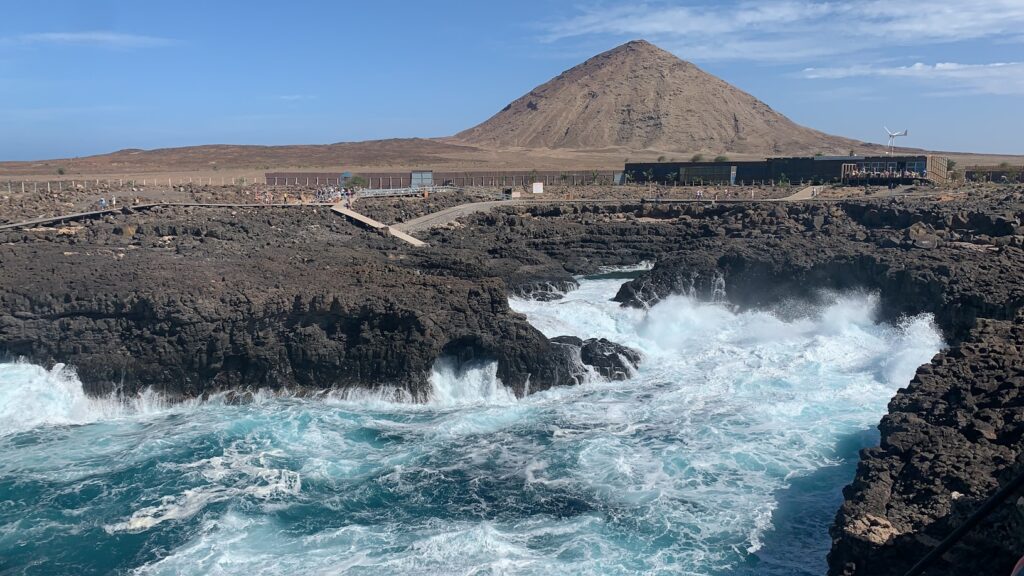 Ondas quebram contra penhascos rochosos escuros com uma montanha seca e marrom e edifícios ao fundo, sob um céu azul claro. Representa eSIM Cabo Verde