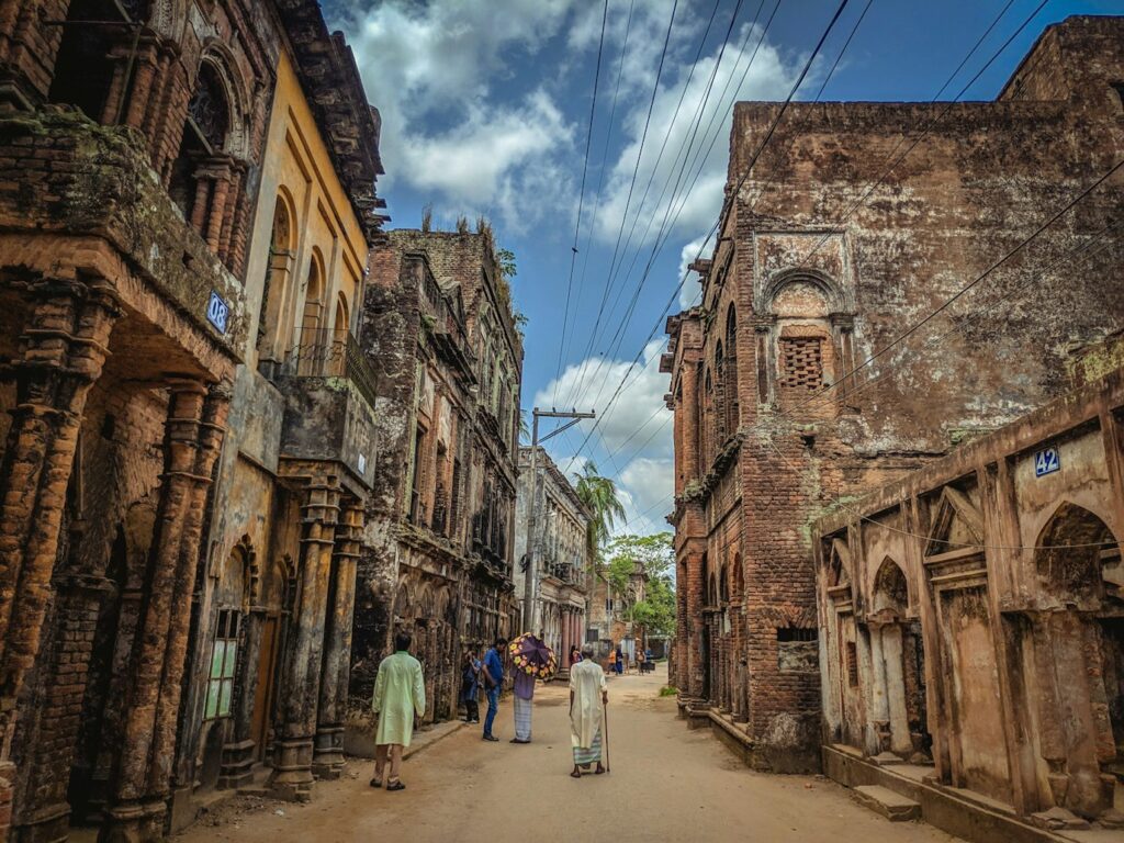 As pessoas caminham por uma rua estreita ladeada por prédios de tijolos antigos e desgastados pelo tempo, sob um céu parcialmente nublado, em Sonargaon. Representa eSIM Bangladesh.