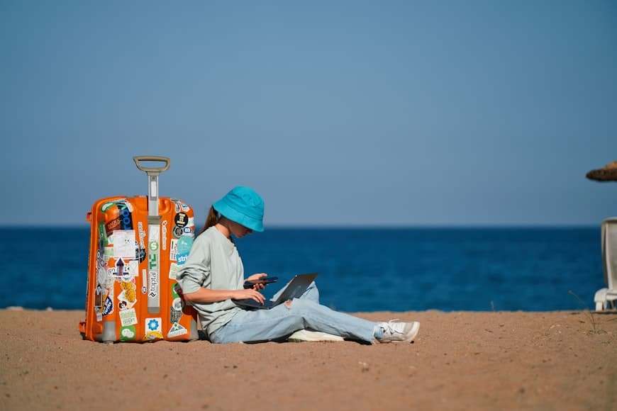 Mulher com chapéu azul sentada na praia usando laptop, com mala de viagem ao lado, apreciando vista do mar e pesquisando no notebook